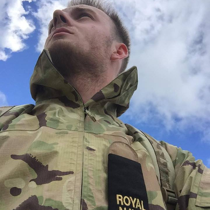 Person wearing a camouflage jacket with a 'Royal Navy' patch against a blue sky.