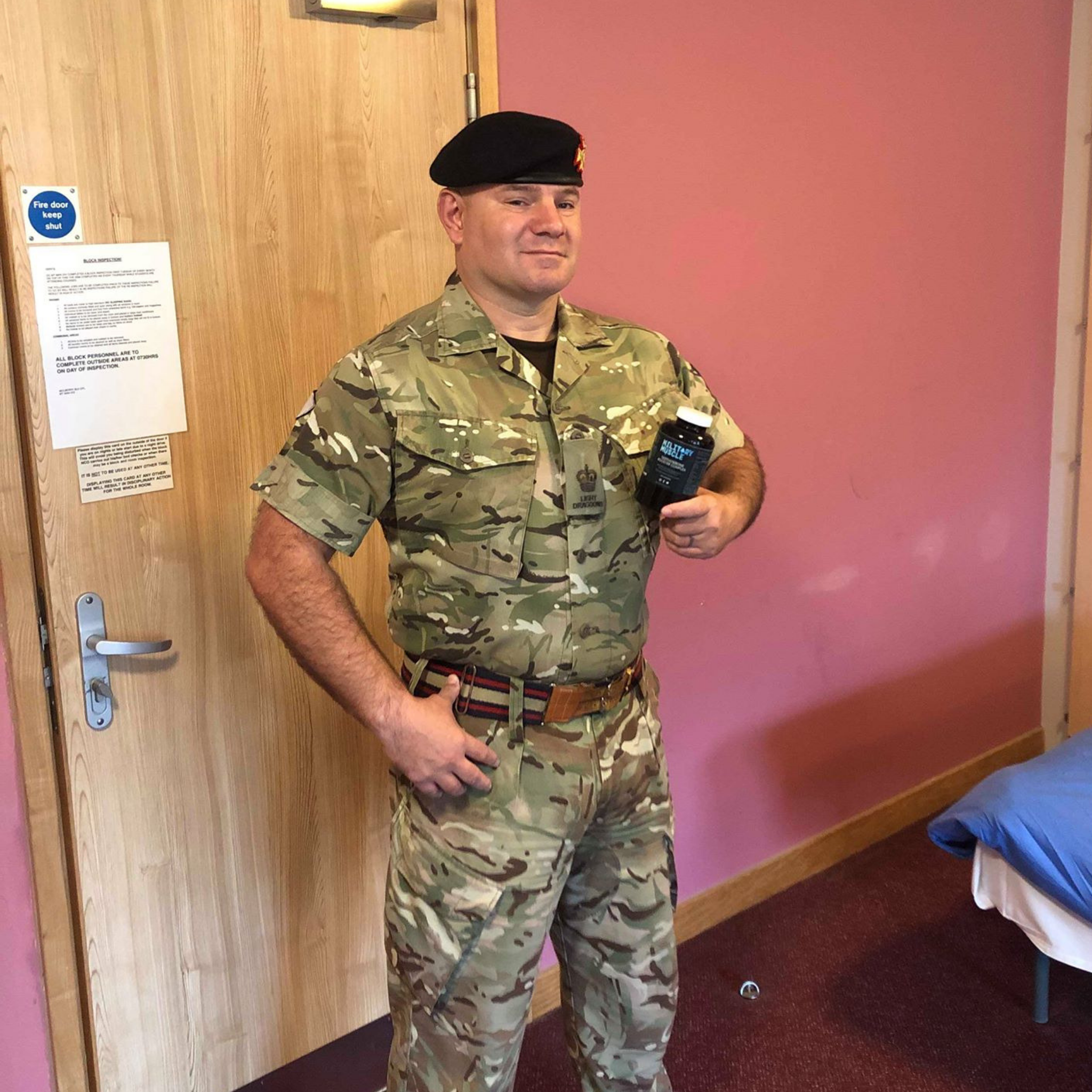 a man wearing full military unifrom holding a package of military muscle testosterone booster whilst atood in front of a wooden door.