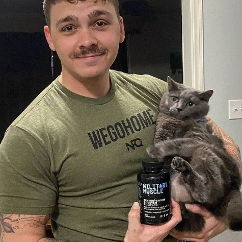 Man holding a cat and a supplement bottle labeled 'Military Muscle' in a casual setting.