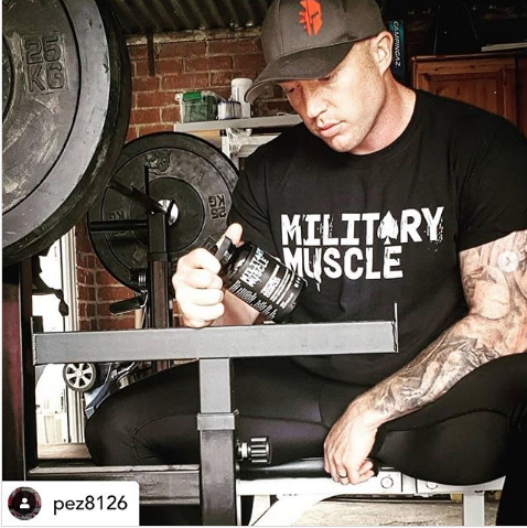 man in a gym setting wearing a black military muscle tee shirt holding a package of military muscle testosterone booster