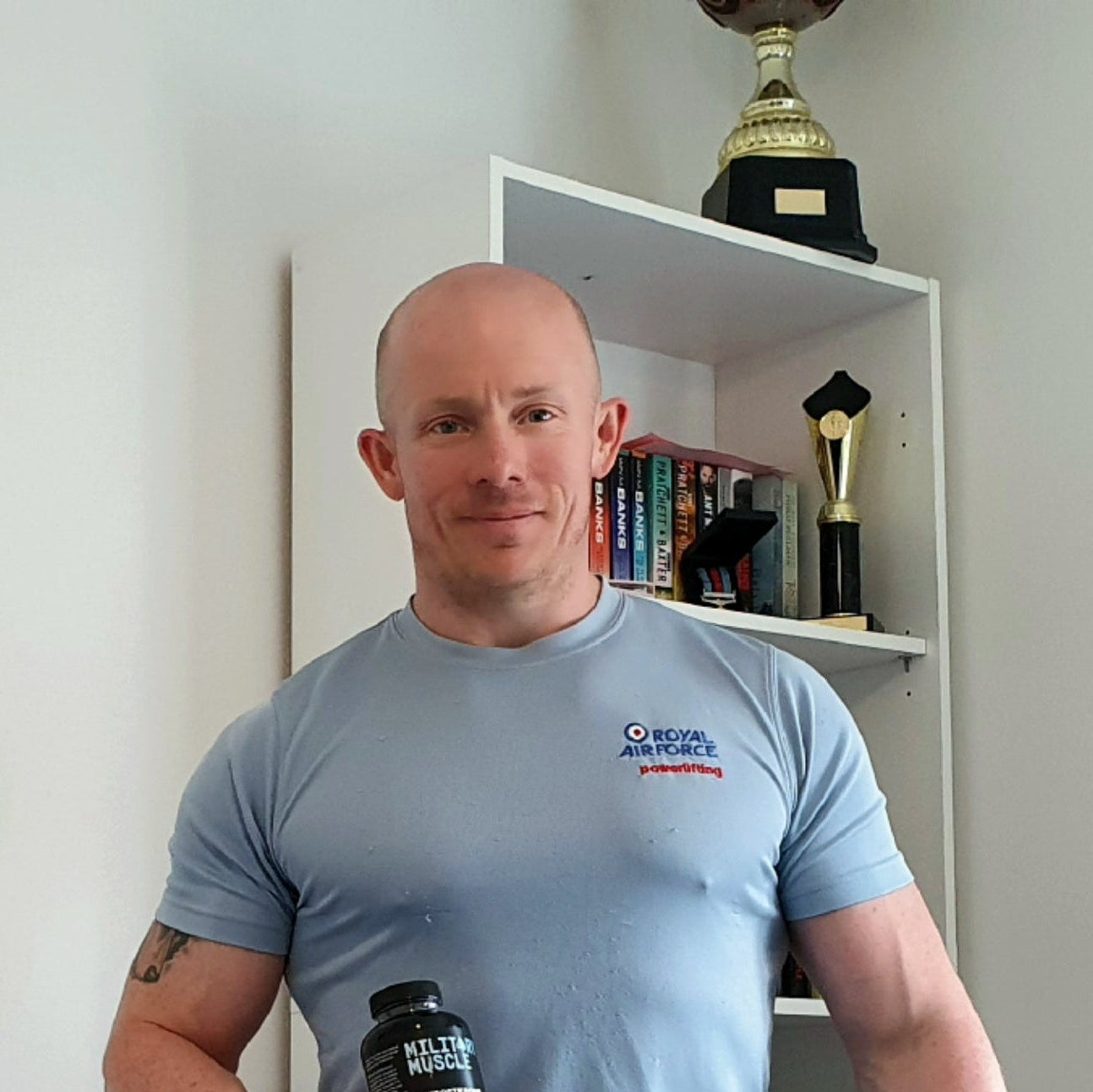 a man wearing a royal air force blue powerlifting tee shirt stood in front of a cabinet with trophies holding a package of military muscle 