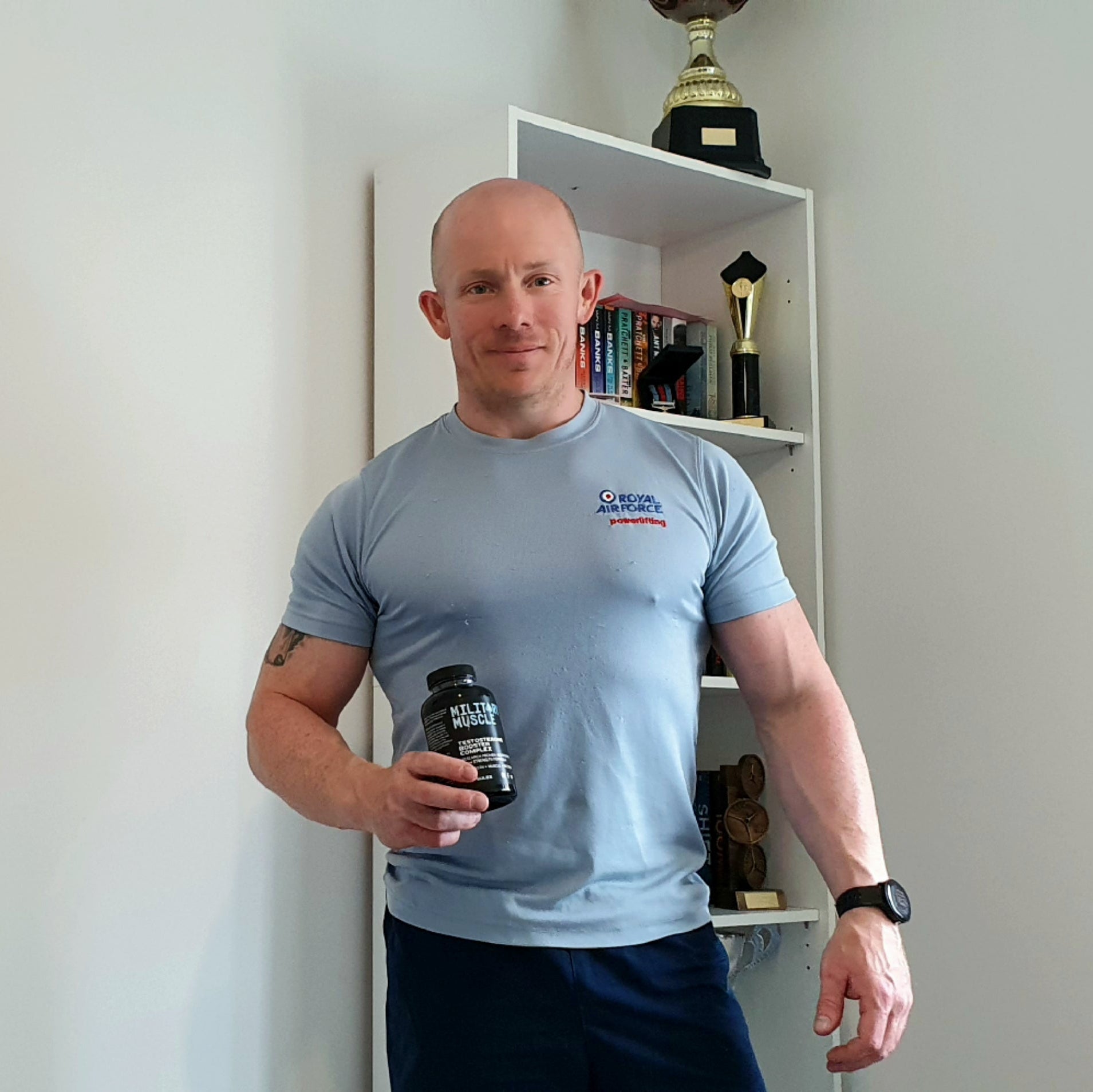 a man wearing a royal air force blue powerlifting tee shirt stood in front of a cabinet with trophies holding a package of military muscle testosterone booster