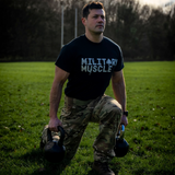 A man on a field wearing a black tee shirt with a military muscle logo and military trousers in the lunge position holding a kettlebell in each hand.