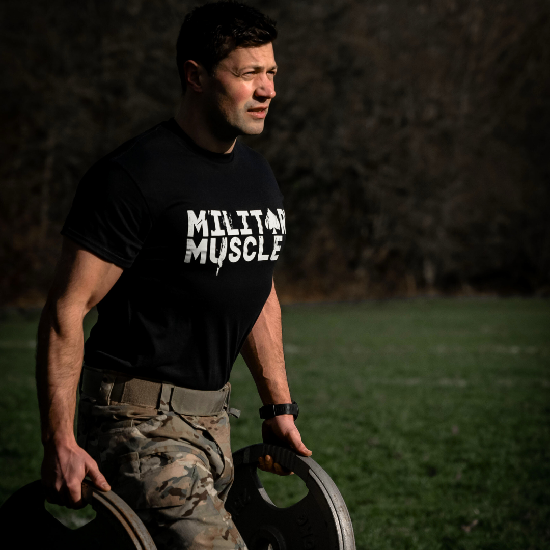 man in a field wearing a black military muscle tee shirt and military uniform trousers carrying a weight plate in each hand.