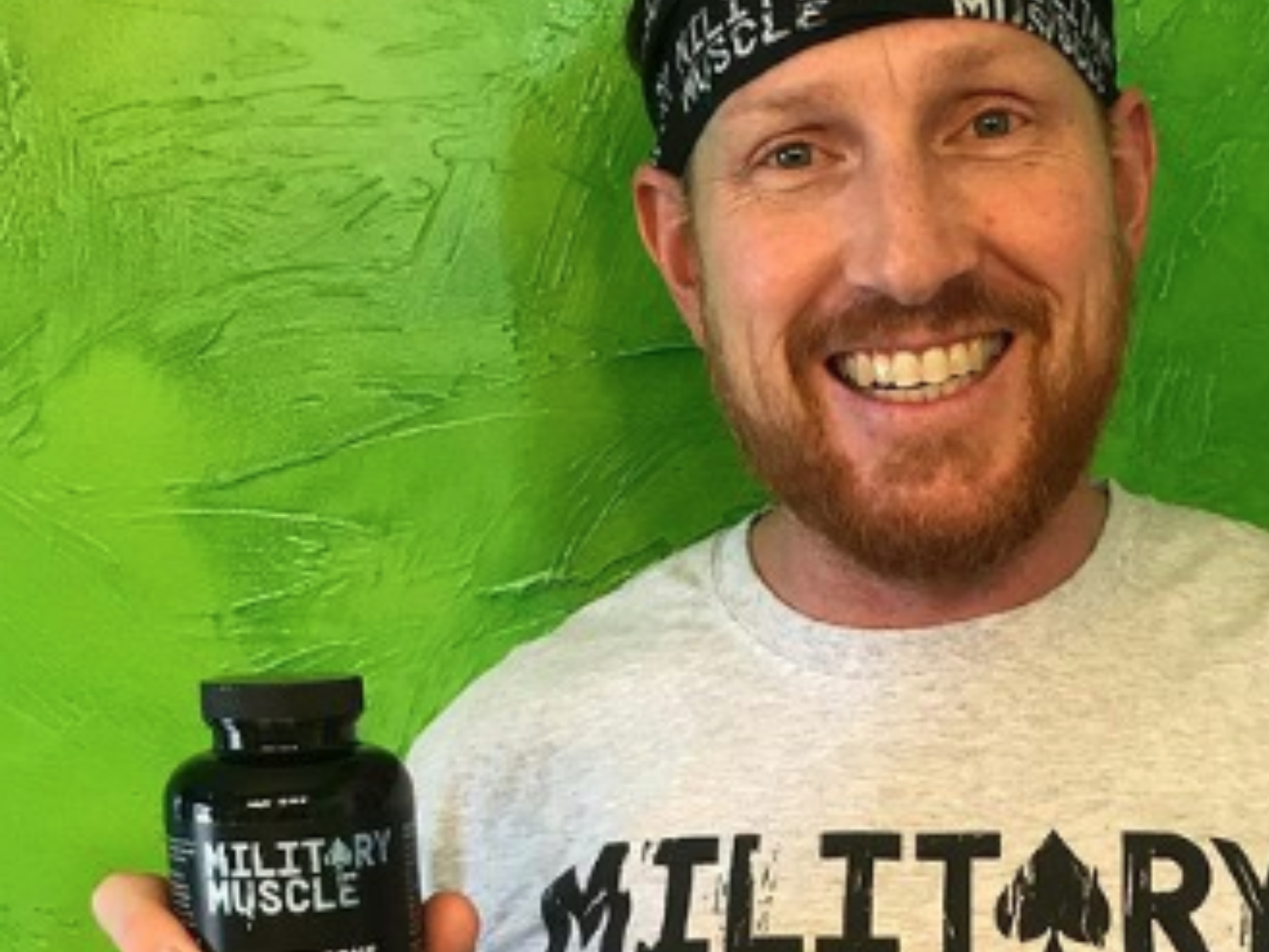 a man standing in front of a green background wearing a military muscle bandana and military muscle tee shirt holding a package of military muscle 